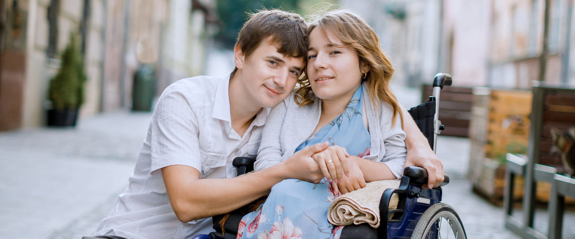 A couple sharing an intimate moment on a cobblestone street - a man crouching beside a woman in a wheelchair, holding hands